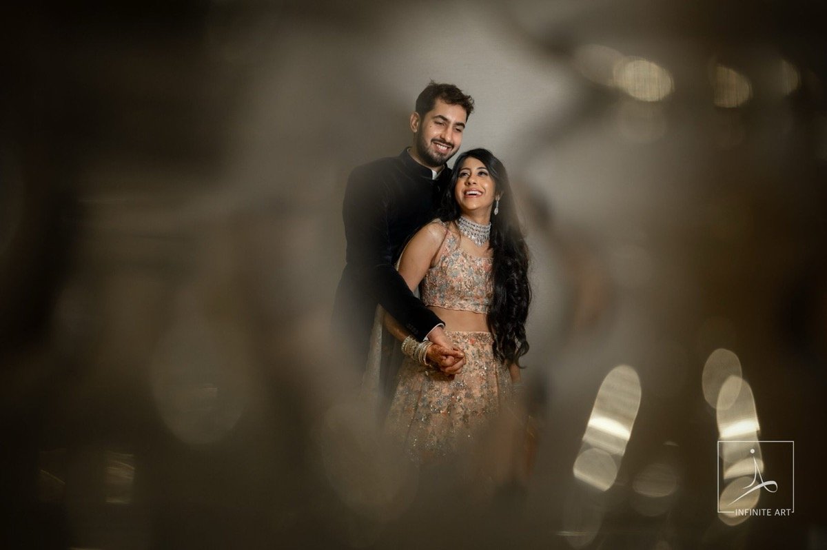 Abhishek and Shweta smiling, framed through soft bokeh — he in a dark bandhgala, she in an embroidered peach lehenga