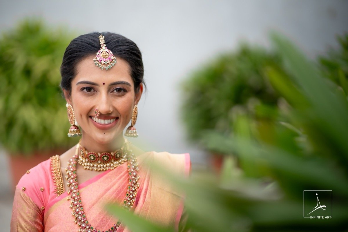 A smiling bride in a pink silk saree, temple jewellery and a maang tikka, framed by soft green leaves