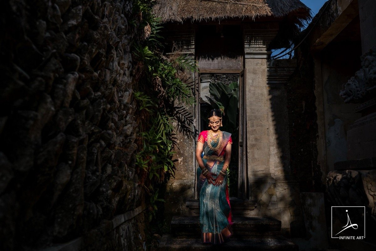A bride in a teal and pink silk saree stands on stone steps in a sunlit doorway beneath a thatched roof