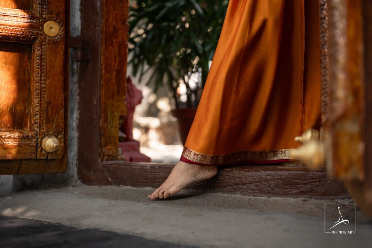 A bride's bare foot steps over a carved wooden threshold, the hem of her orange silk saree lifted, in warm afternoon light